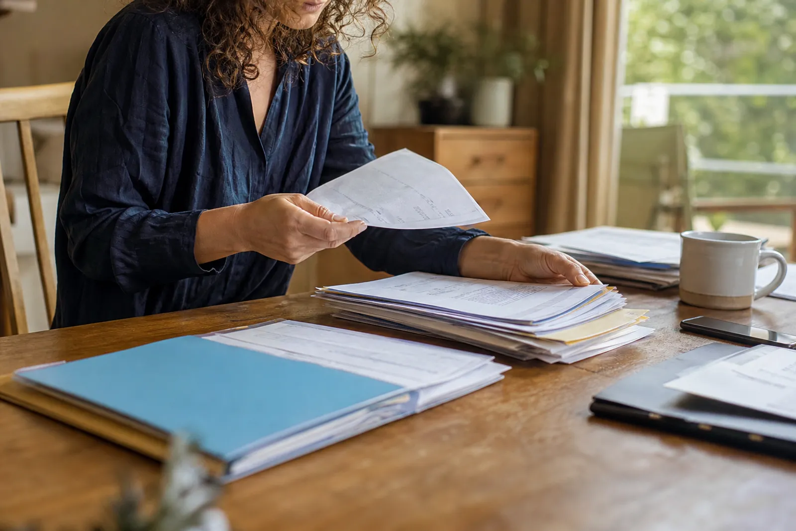 Une personne trie des documents administratifs et des factures dans des chemises cartonnées sur une table en bois.