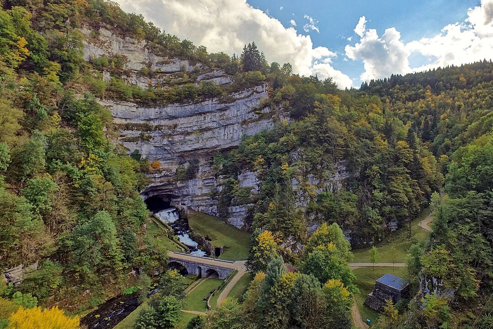 Un voyageur marche au bord d’une rivière jurassienne, avec en arrière-plan des falaises calcaires et une ville historique au loin.